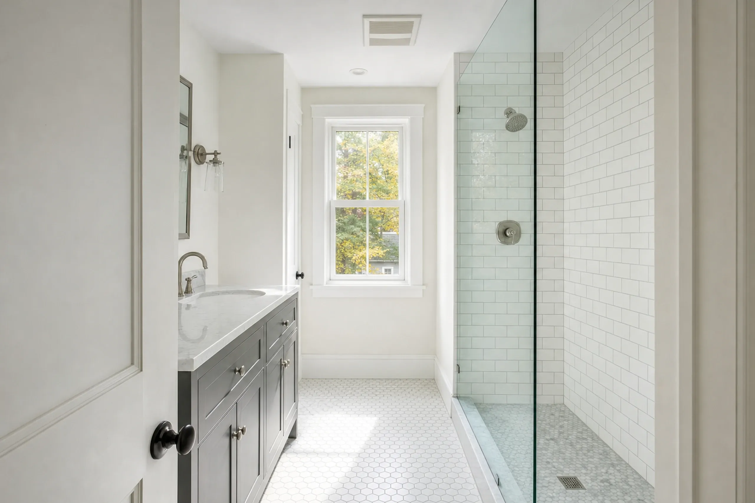 Bathroom remodeling: narrow bathroom with a gray vanity and marble top, a glass-walled subway tile shower, white hexagon floor tile and a window onto trees