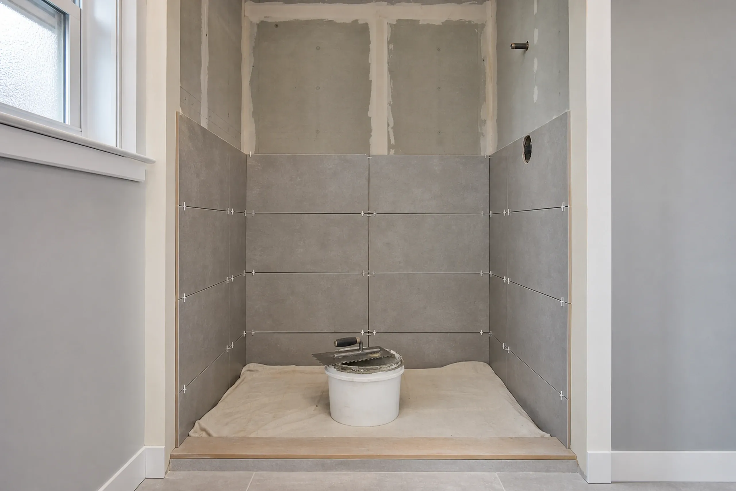 Bathroom remodeling: shower wall half set in large gray porcelain tiles with spacers in the joints, a bucket of thinset and a trowel on a drop cloth