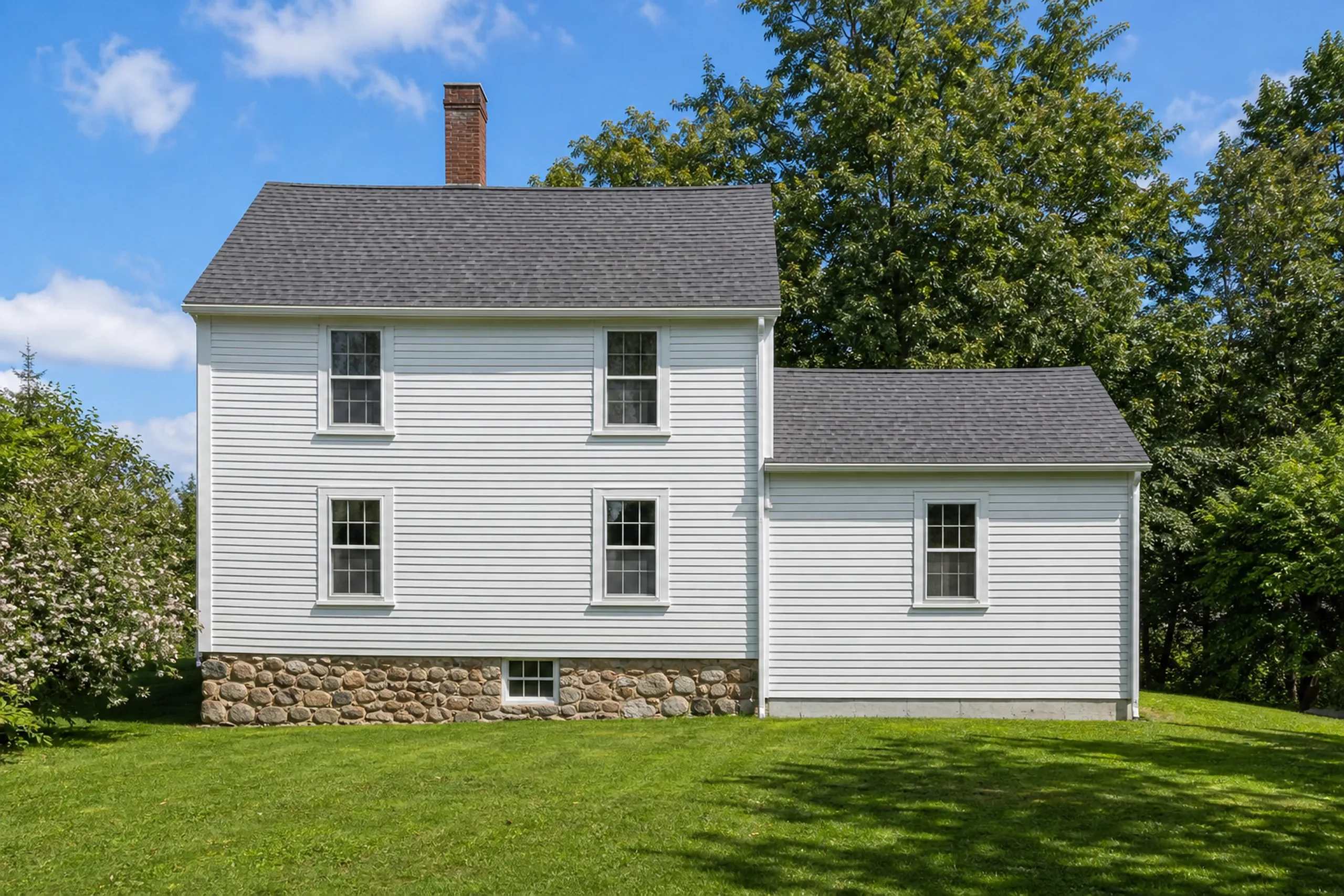 General contractor in Biddeford: two-story white clapboard house with a lower side wing, a brick chimney and a fieldstone foundation, on a lawn in front of tall trees
