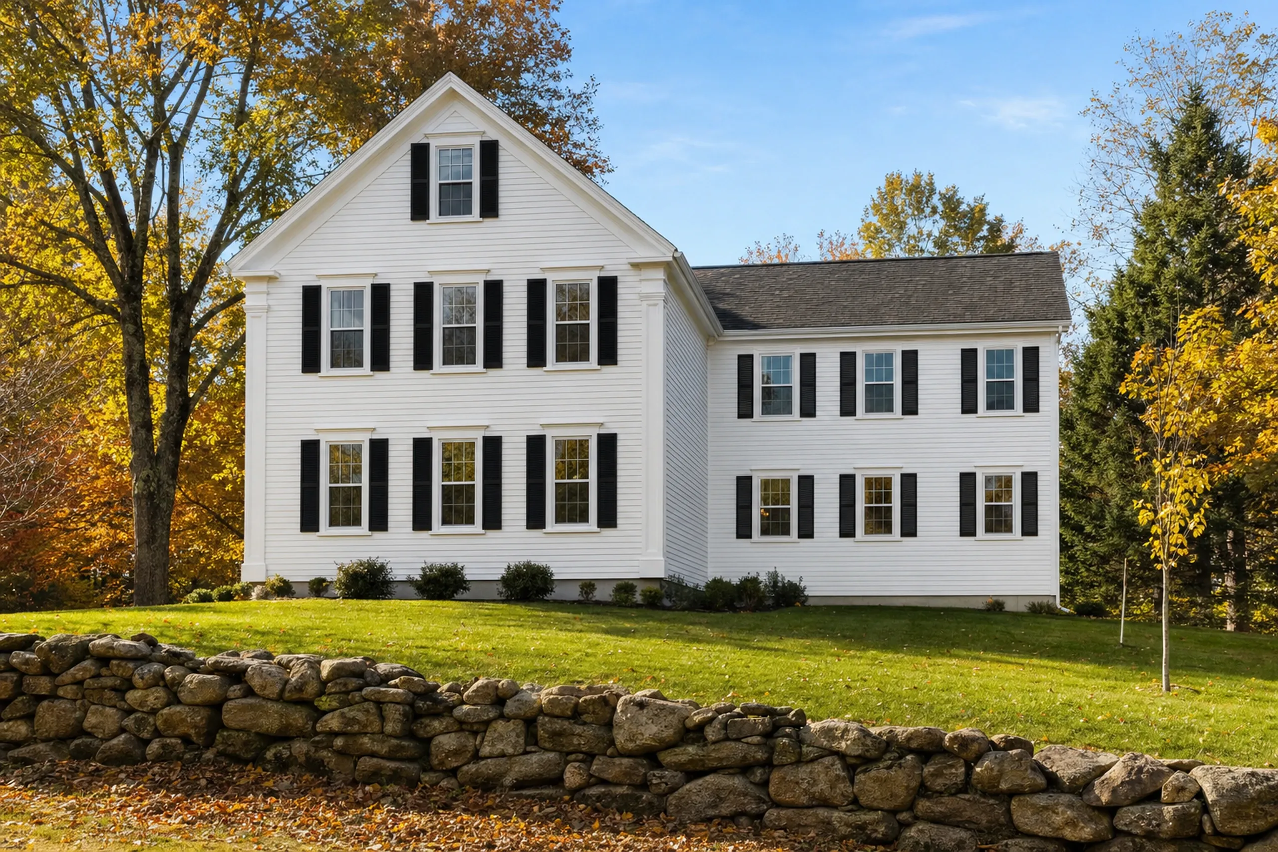 General contractor in Freeport, ME: A white two-story house with black shutters and a lower side wing, set behind a fieldstone wall and lawn in autumn