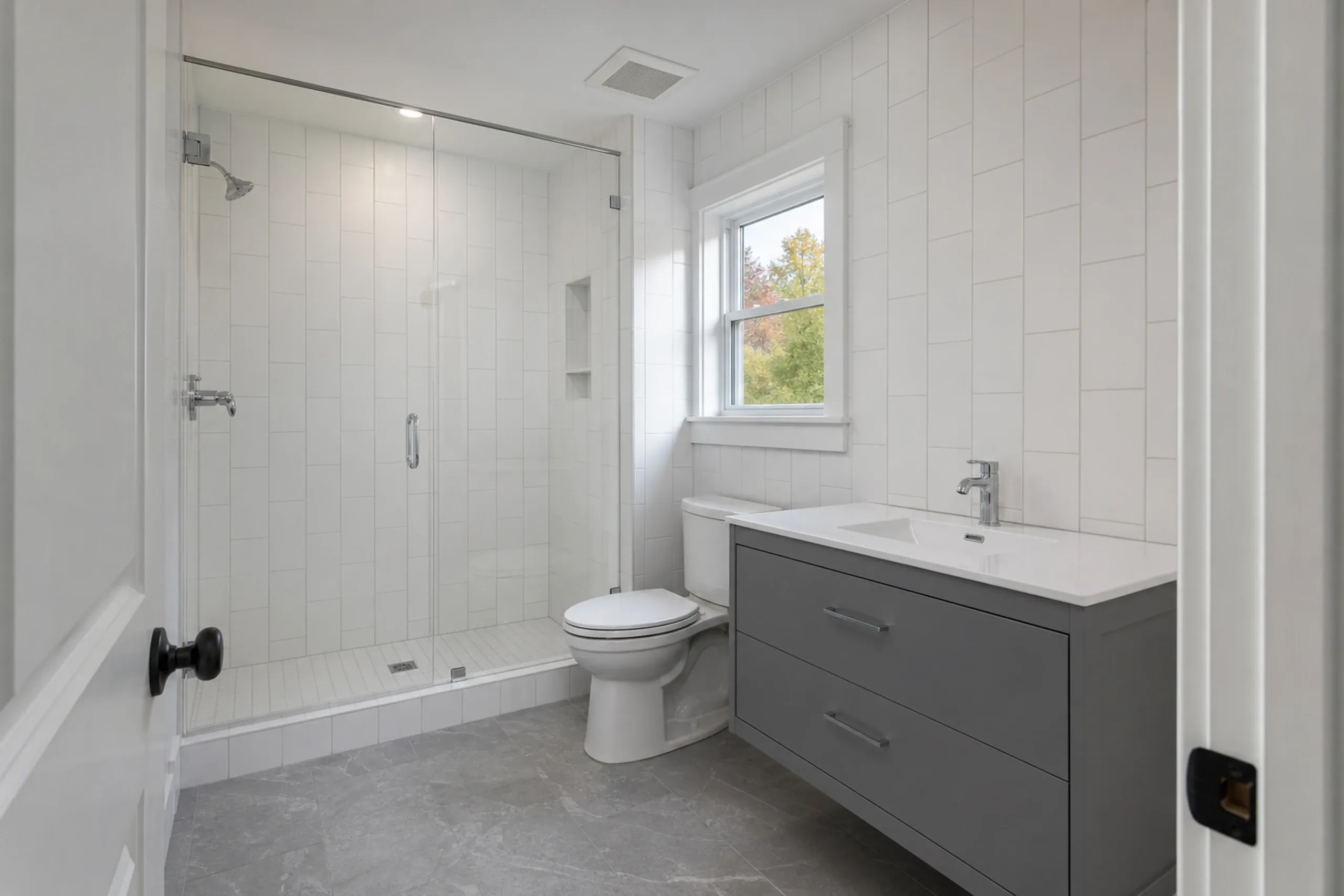 General contractor in Gorham: A bathroom with a glass-door tiled shower, white vertical wall tile, a white toilet and a gray wall-hung vanity beneath a window