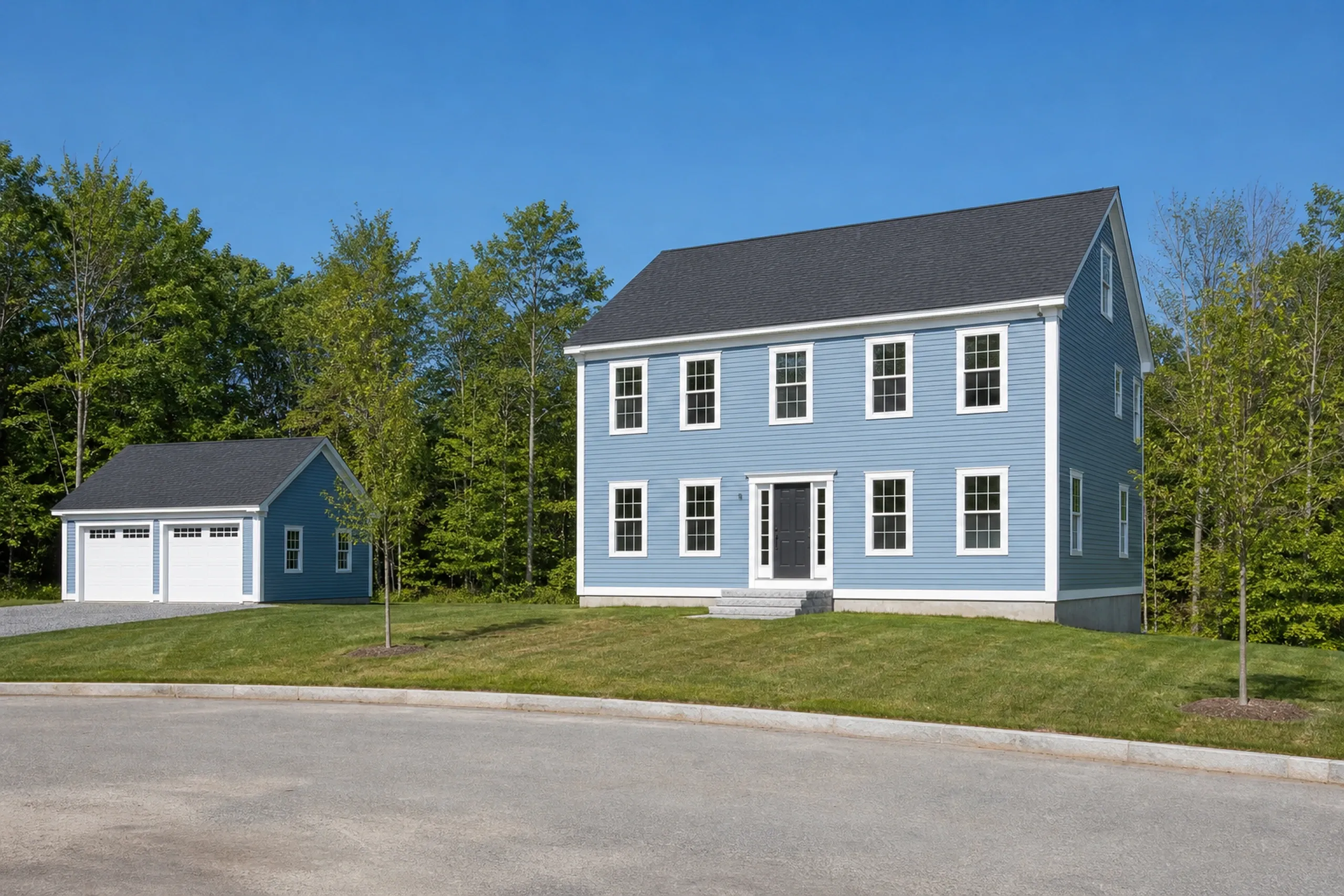 General contractor in Scarborough: blue clapboard two-story house with a dark roof and gray front door, a detached two-door garage and a curved paved street with a young tree