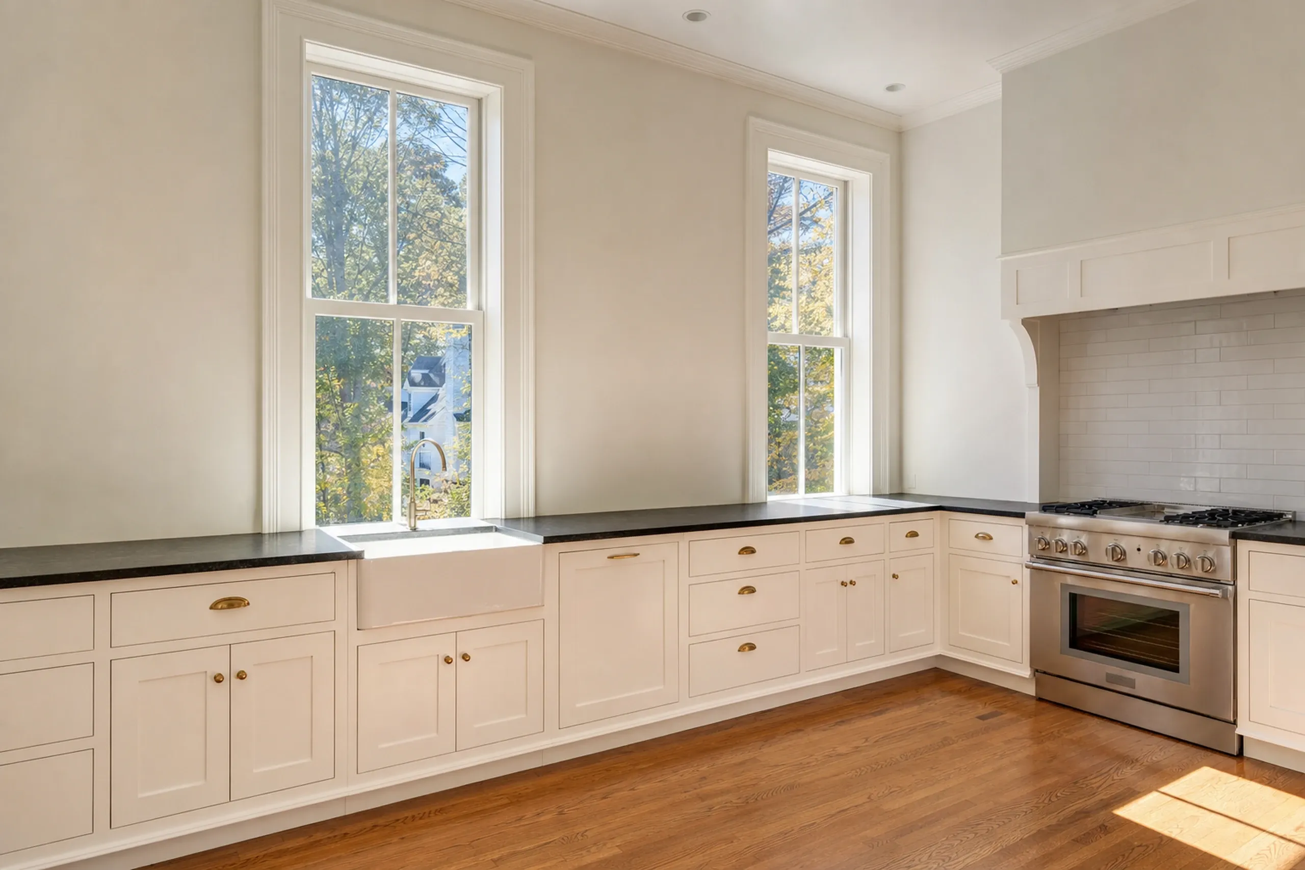 Kitchen remodeling: tall double-hung windows above a farmhouse sink, with cream cabinets, dark counters, brass pulls, white tile and a range at right