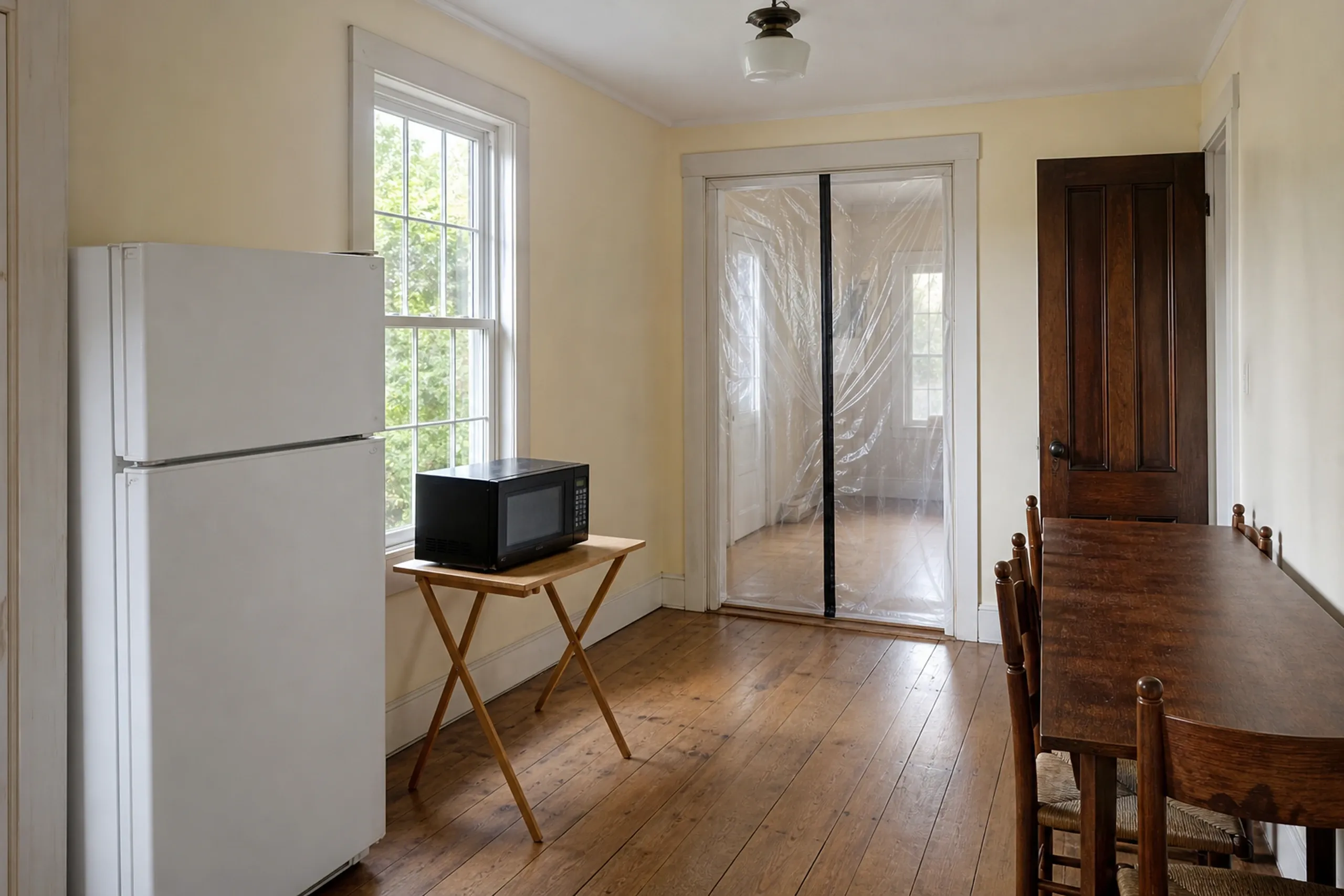 Kitchen remodeling: white refrigerator and a microwave on a folding tray table in a room with plank floors, a wood table and plastic sheeting in the doorway